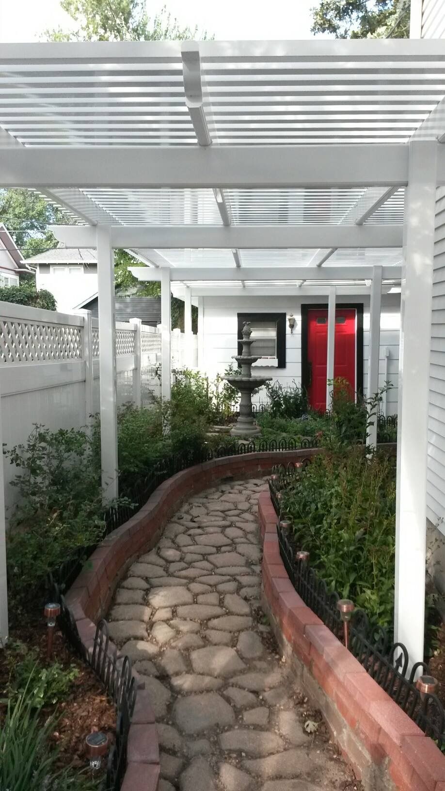 Stone pathway curves through a garden, leading to a white building with a red door, under a white pergola.