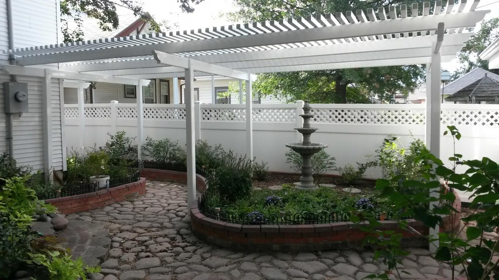 White pergola over a stone patio with a fountain and garden beds, enclosed by a white fence.
