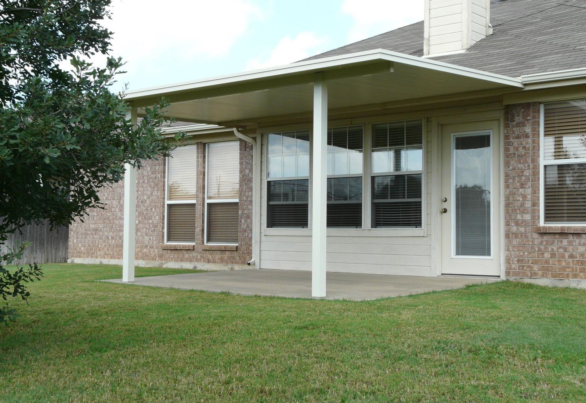 Patio with white columns, tan roof, and brick house. Green grass in foreground.