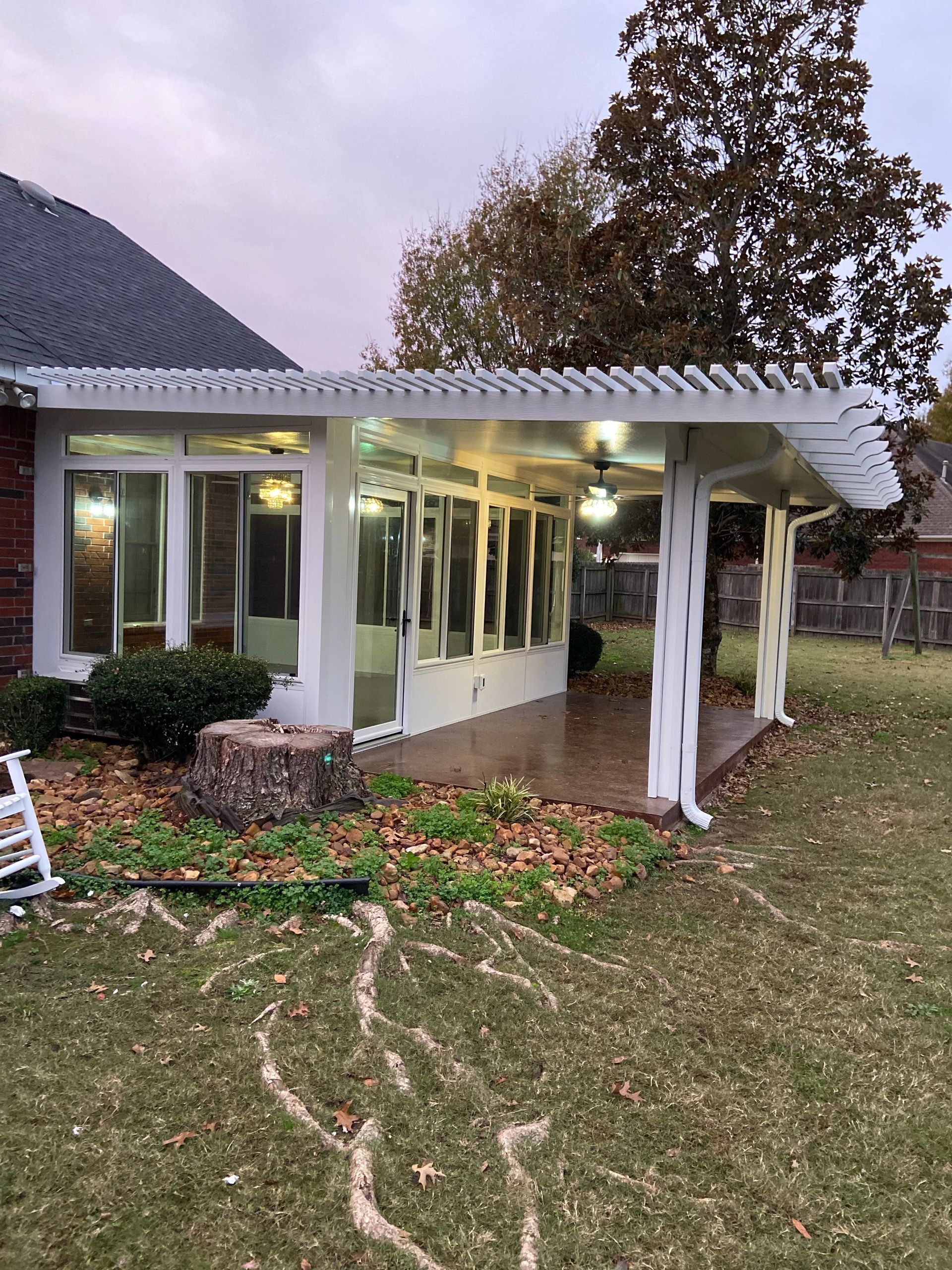 White-framed sunroom with pergola roof; brown concrete floor; green lawn with leaves; overcast sky.