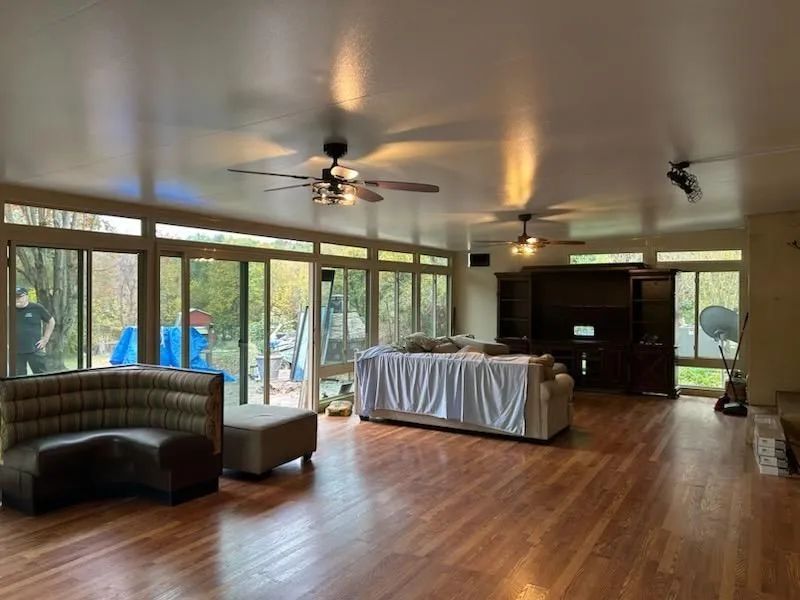 Spacious room with wooden floors, large windows, ceiling fans, furniture covered in white, and a sec