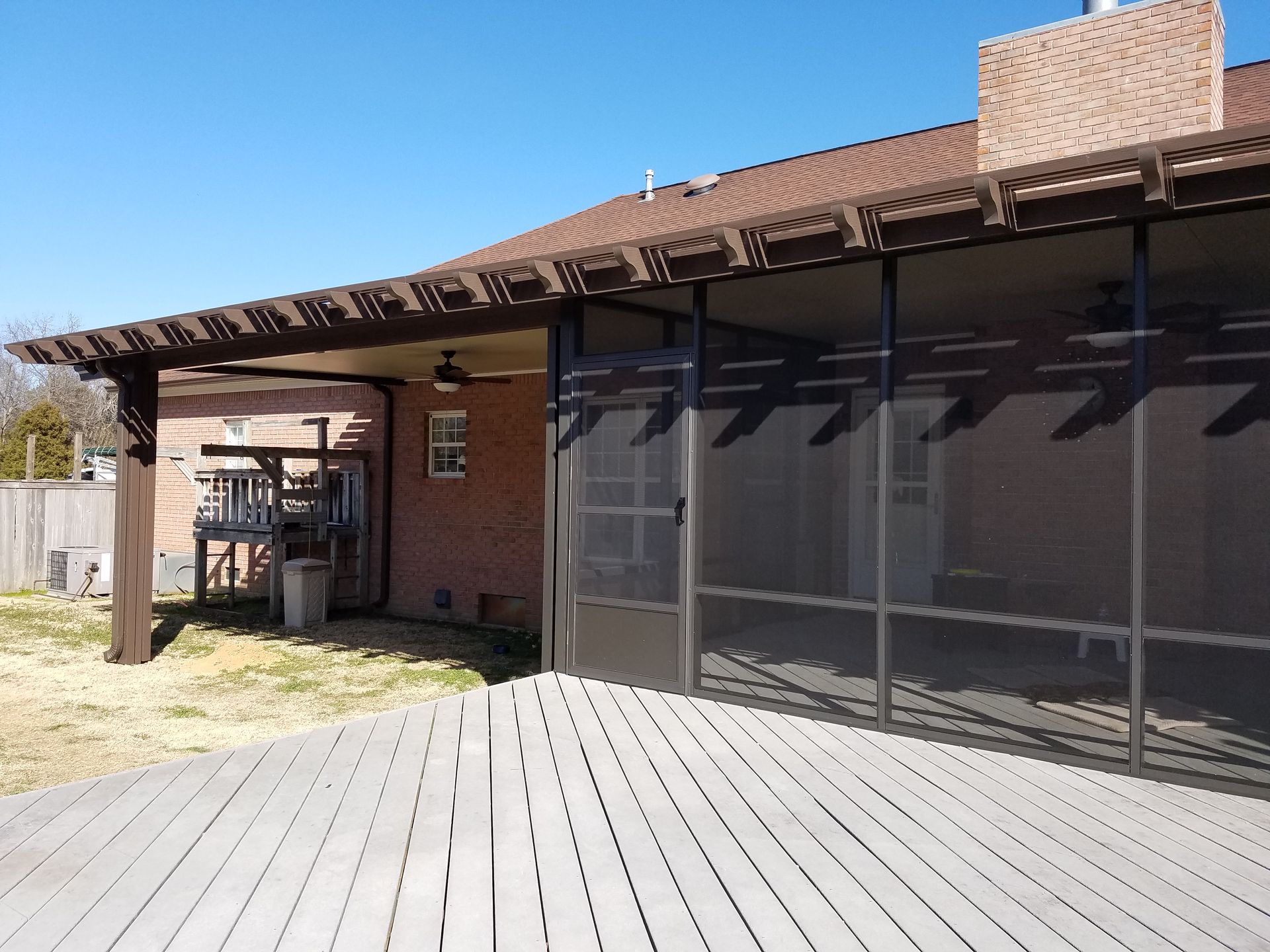 Wooden deck leads to a screened-in porch with a pergola. Brick house and blue sky in the background.