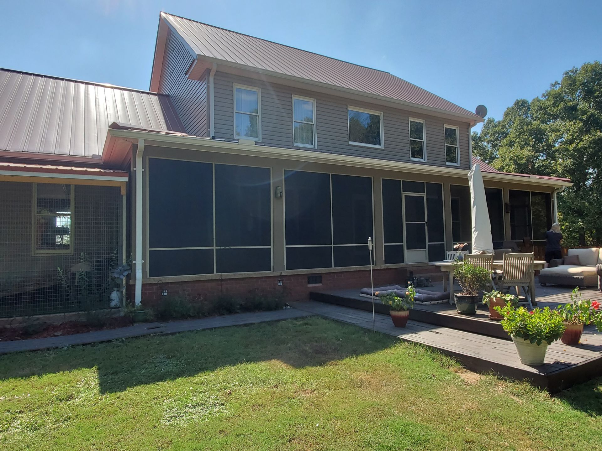 Two-story house with a screened porch and metal roof on a sunny day. Green grass and potted plants in the yard.