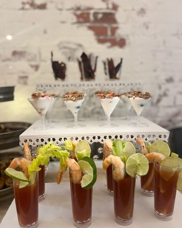 Food display with Bloody Marys, shrimp, parfaits, and chocolate bark. Against brick wall.