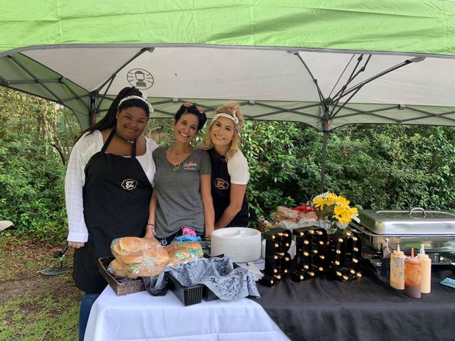 Three women stand behind a food table at an outdoor event. They smile, wearing aprons and headbands.