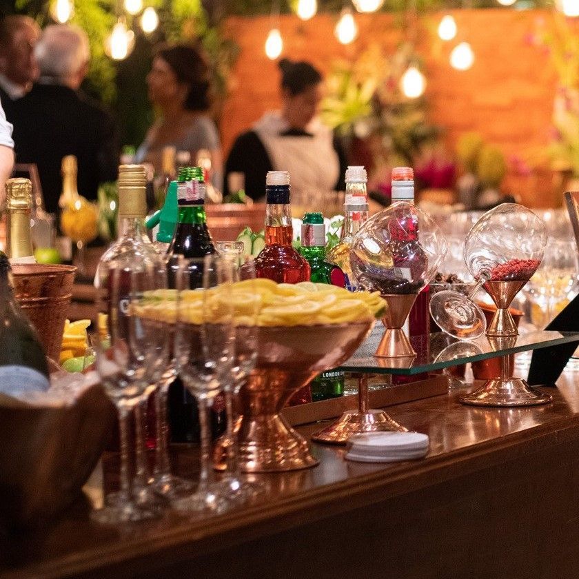 Cocktail bar with bottles, glassware, snacks, and people in the background, lit by soft lighting.