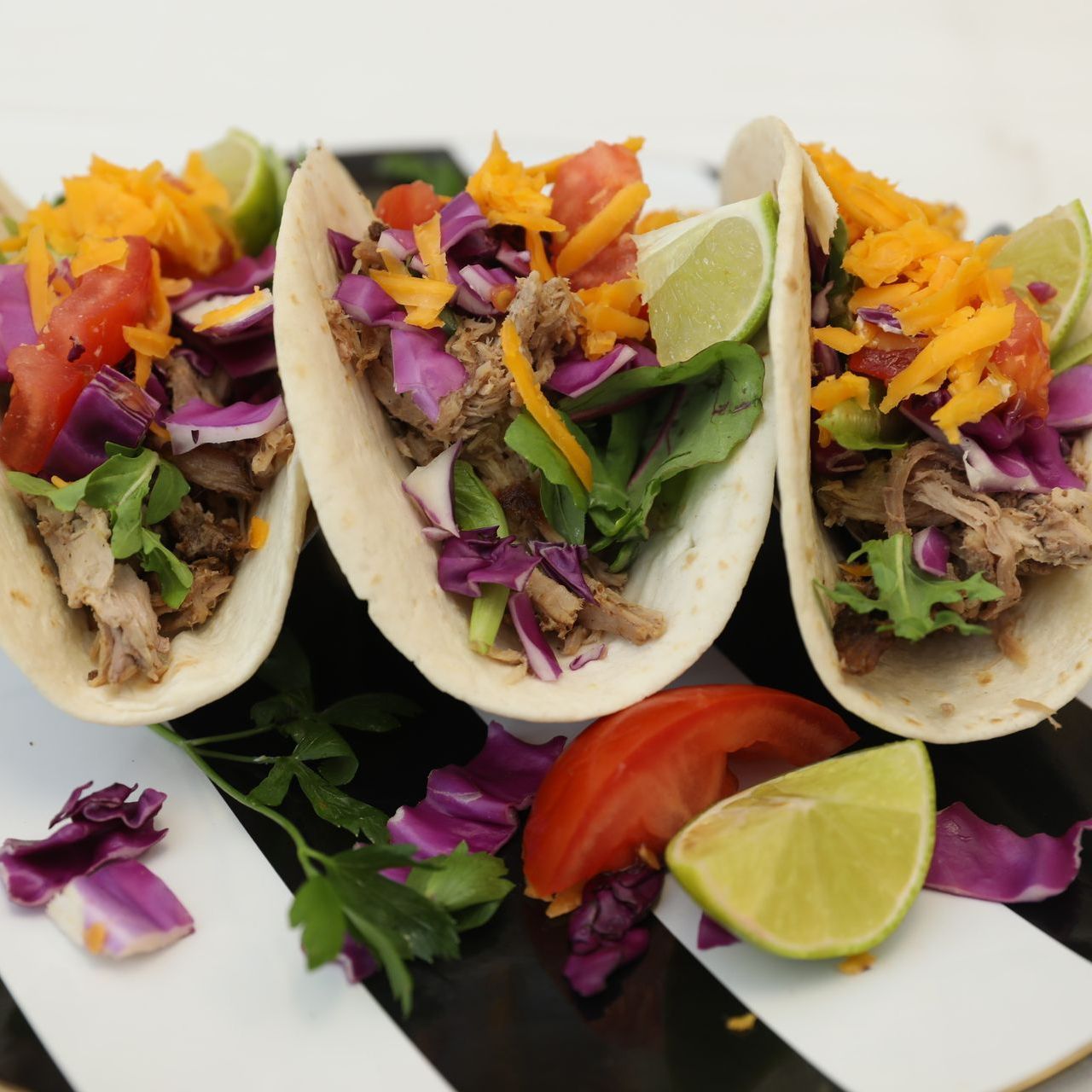 Three tacos filled with meat, vegetables, and cheese, garnished with lime and served on a striped plate.