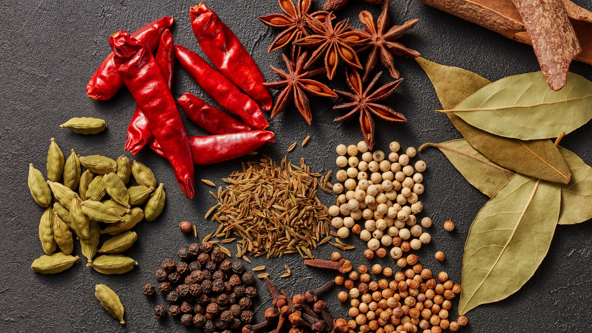 Assorted spices: red chilies, star anise, cardamom, peppercorns, cinnamon sticks, bay leaves on a dark surface.