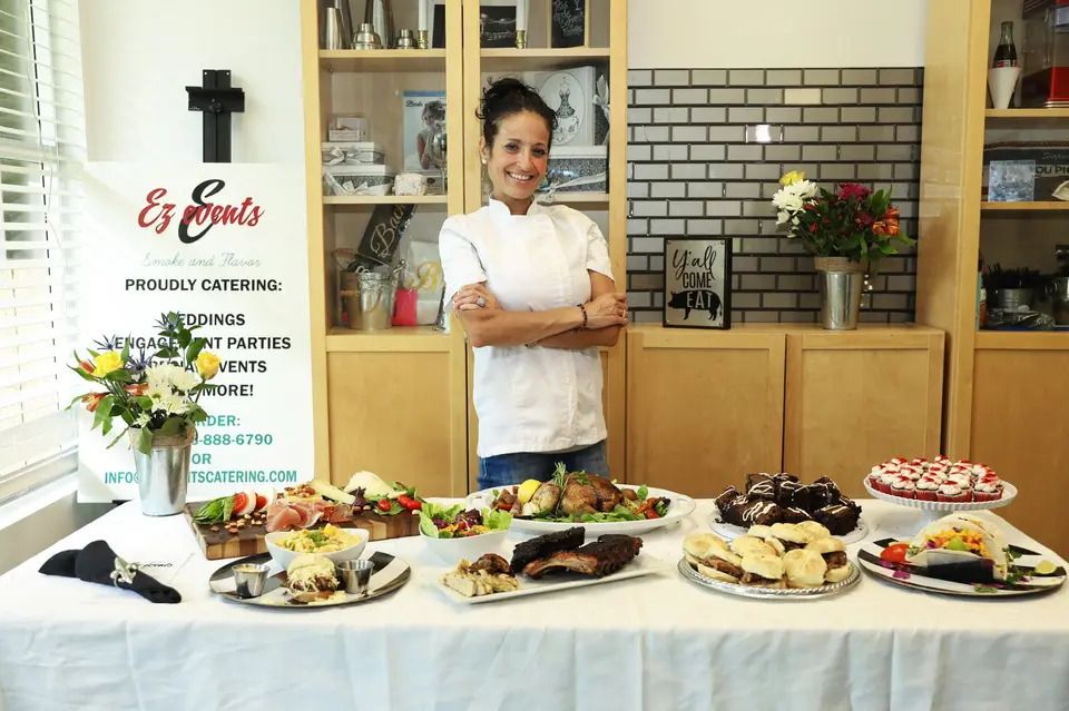 Woman chef stands behind a table of catered food, smiling with arms crossed.