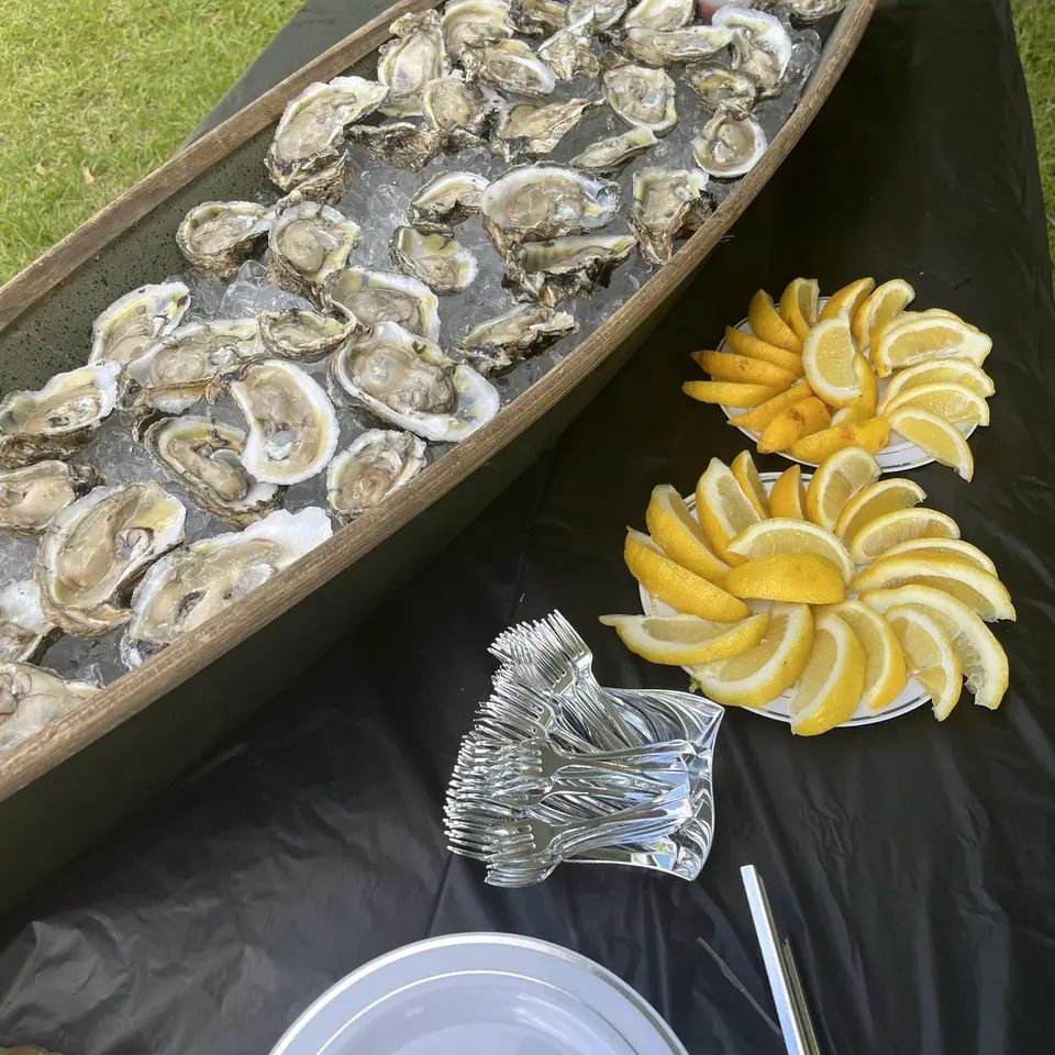 Oysters on ice in boat-shaped container with lemon wedges and forks on black tablecloth outdoors.