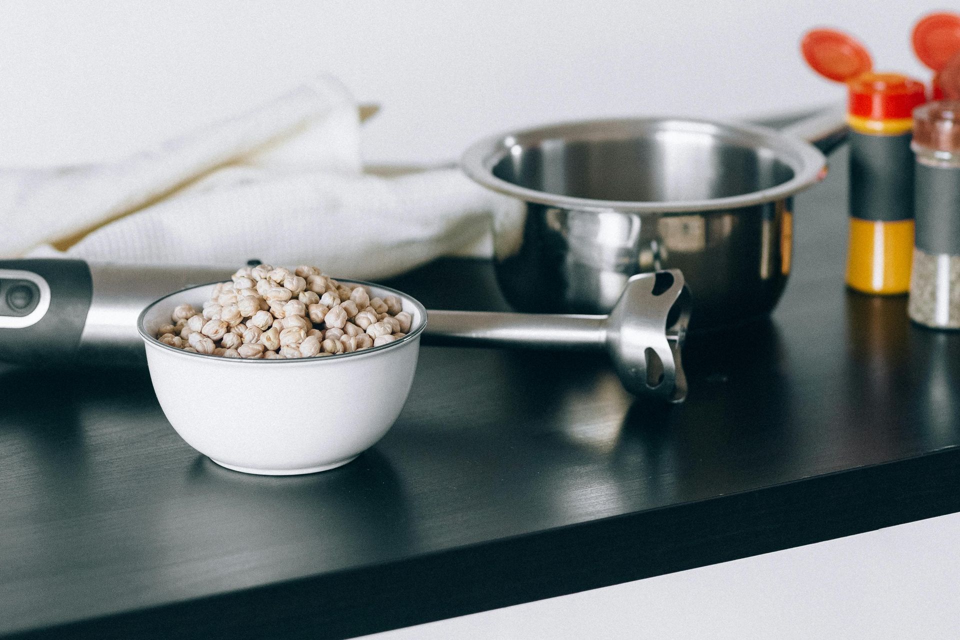 White bowl with brown grains, immersion blender, pot, spices on black surface.