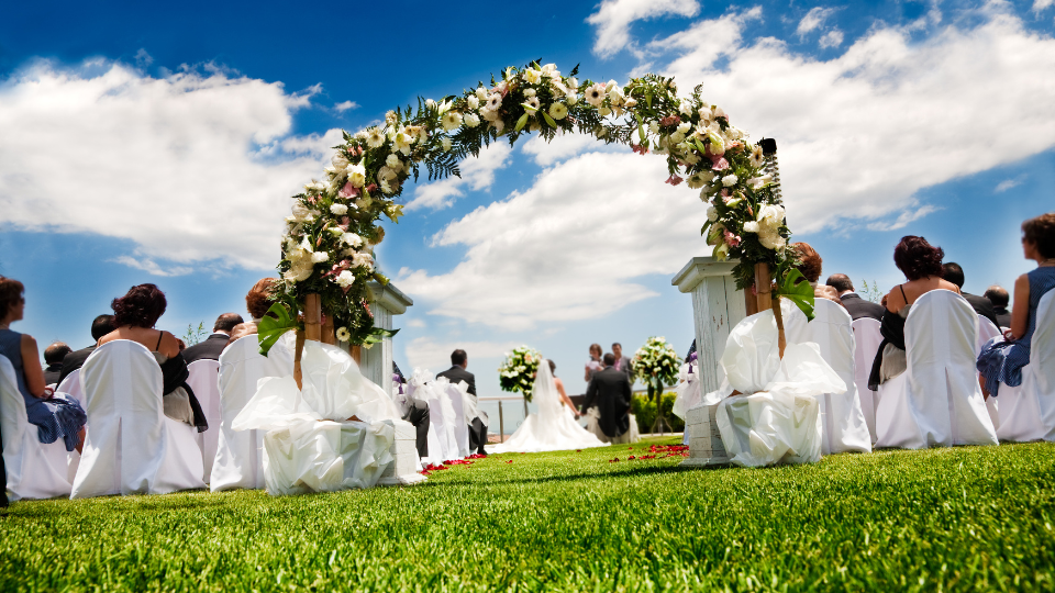Wedding ceremony outdoors, floral archway, bride and groom, guests seated, sunny day.
