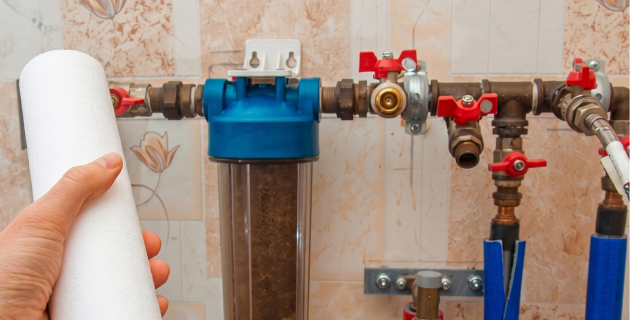 Hand holding a white filter cartridge beside a blue water filter and plumbing pipes under a sink