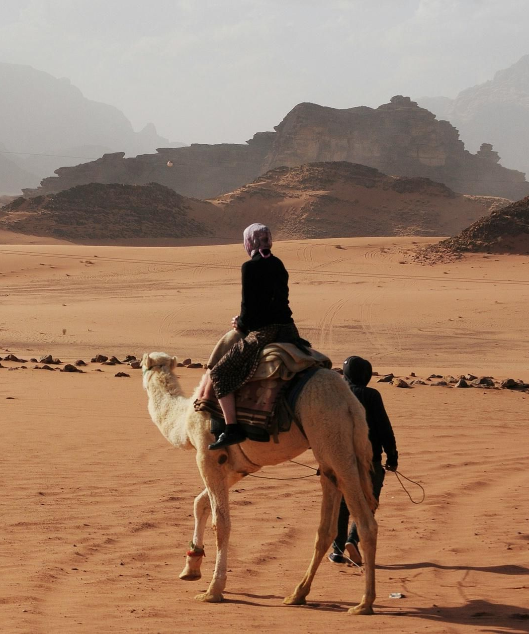 Person rides a camel across a sandy desert with rocky hills in the background.