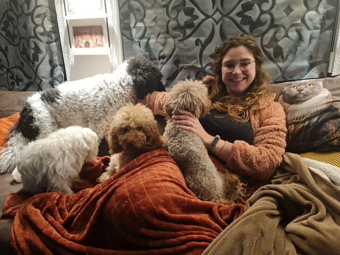 Een vrouw zit lachend op een bank, met meerdere honden die onder dekens liggen te knuffelen.