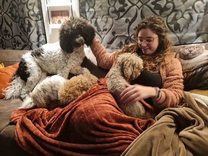 Vrouw ligt languit op een bank met drie honden met krullend haar, lachend en één van de honden aaiend.