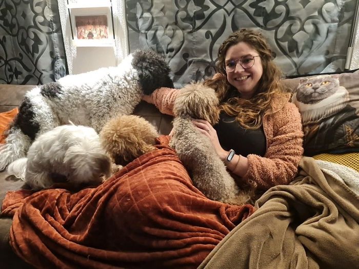 Een vrouw zit lachend op een bank, omringd door verschillende pluizige honden en dekens.