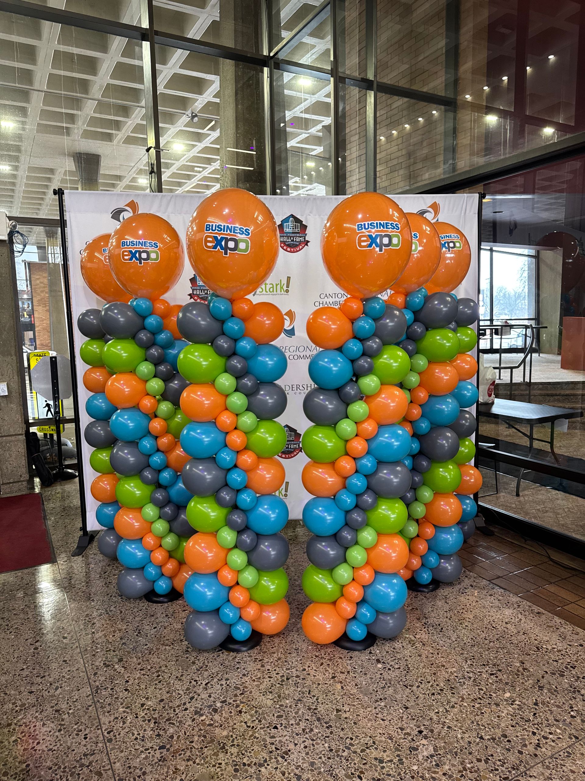 Colorful flat balloon columns with branded orange “Business Expo” balloons displayed in front of a step-and-repeat backdrop for a corporate event by Designs by Jo Jo’s Creations in Canton, Ohio.