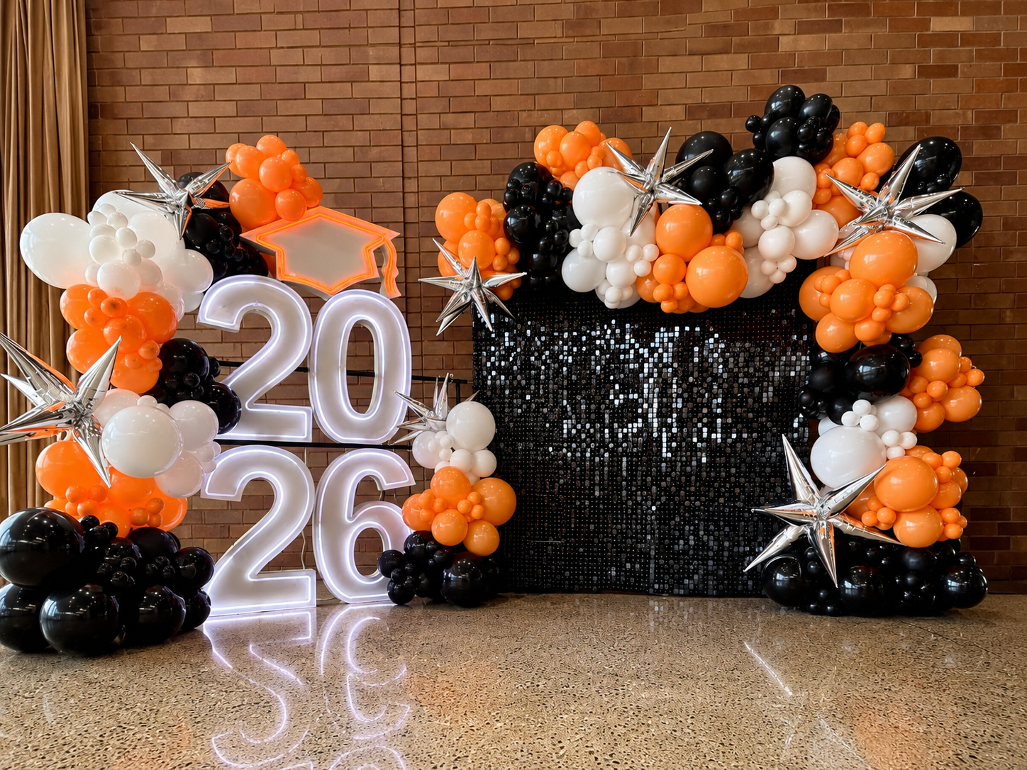 Stacked illuminated “2026” marquee numbers with a graduation cap, black shimmer wall, and orange, black, and white balloon garlands.