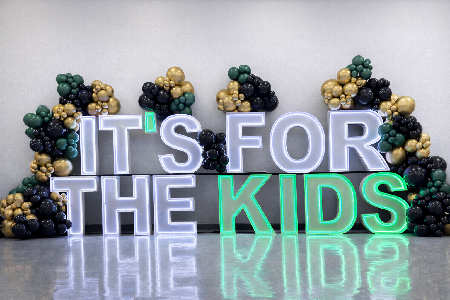 Illuminated “IT’S FOR THE KIDS” marquee letters with black, metallic gold, and dark green balloon accents.