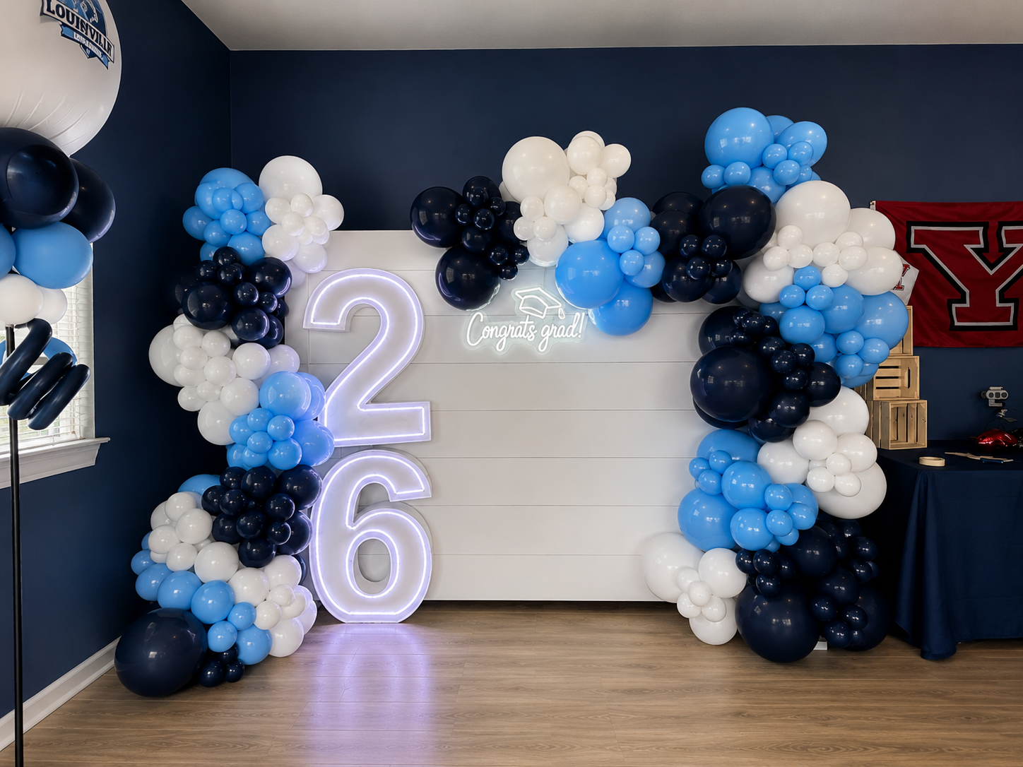 Stacked “26” LED marquee numbers beside a white backdrop with a “Congrats Grad!” neon sign and light blue, navy, and white balloon garland.