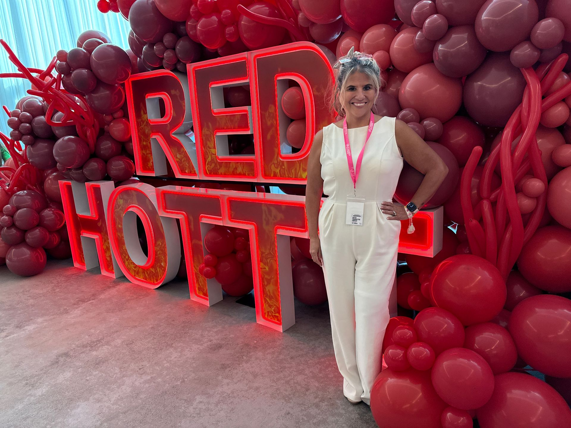 Jo Jo Schneider, owner of Designs by Jo Jo’s Creations, posing beside red LED marquee letters spelling “RED HOT” and a large red and burgundy balloon installation.