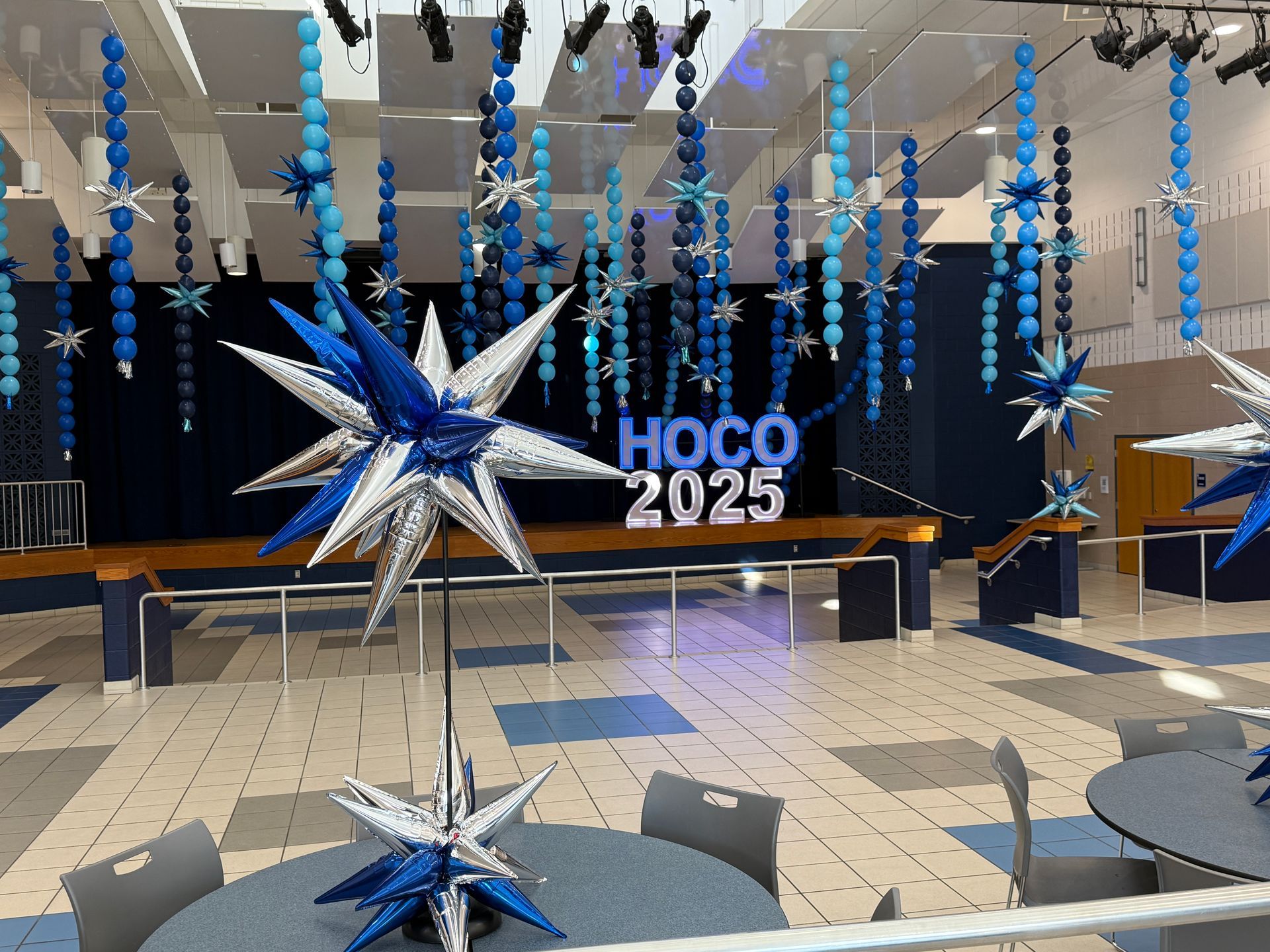 Blue and silver suspended balloon strands with metallic starbursts, table centerpieces, and illuminated “HOCO 2025” marquee letters.