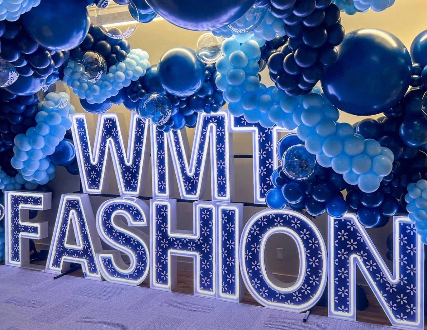 Custom illuminated “WITH FASHION” marquee letters surrounded by light blue, royal blue, and navy balloon décor with clear bubble balloons.