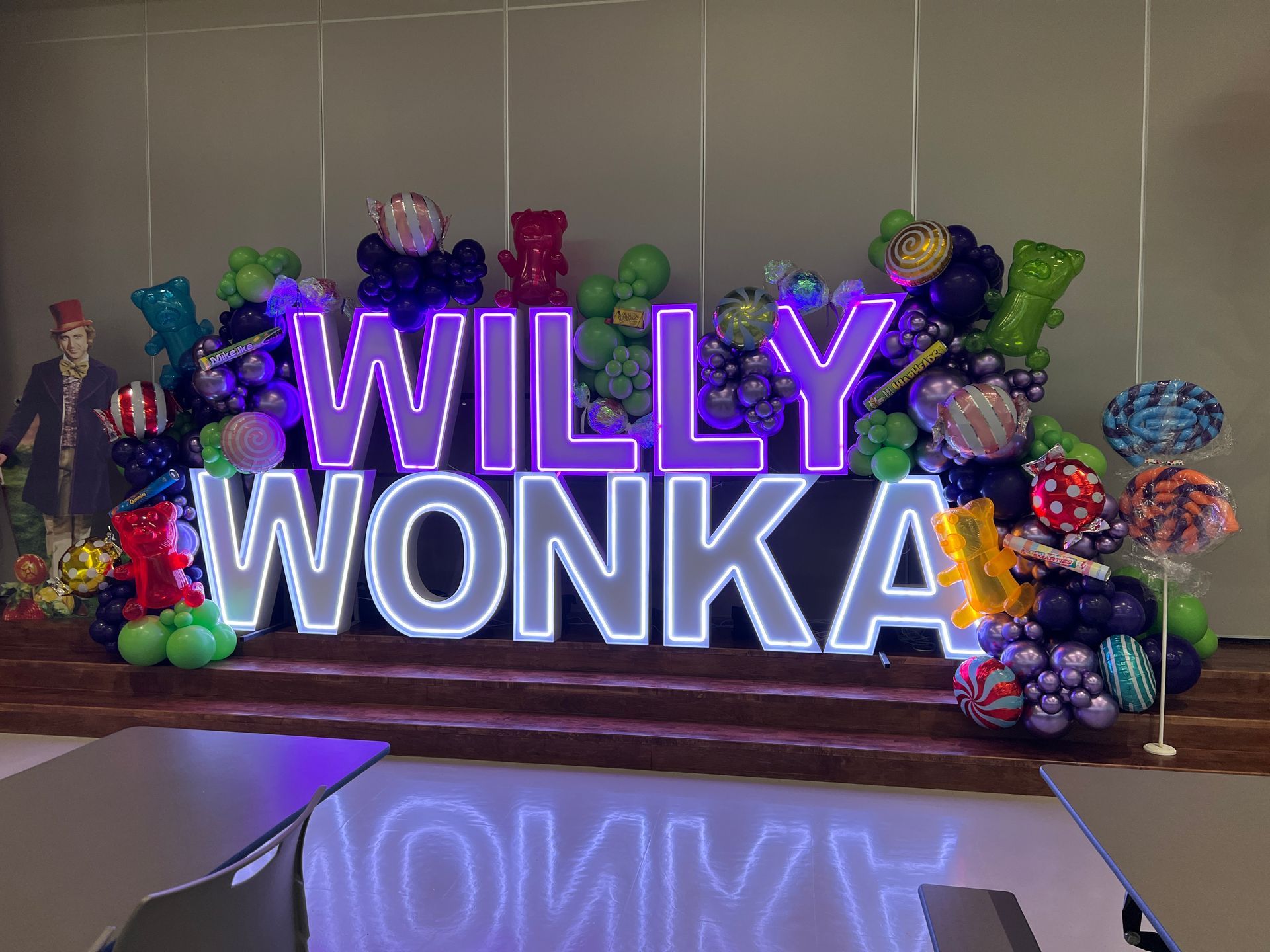 Illuminated “WILLY WONKA” marquee letters surrounded by purple, green, and silver balloons with oversized candy and gummy bear decorations.