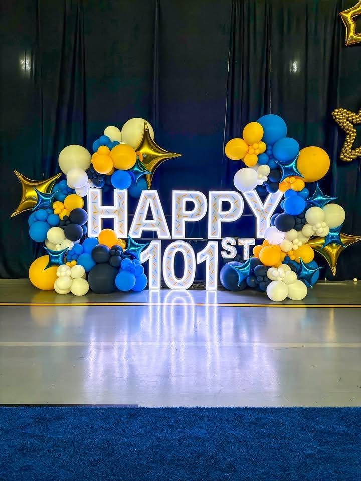 Illuminated “HAPPY 101ST” marquee display surrounded by blue, gold, white, and navy balloons with metallic star accents.