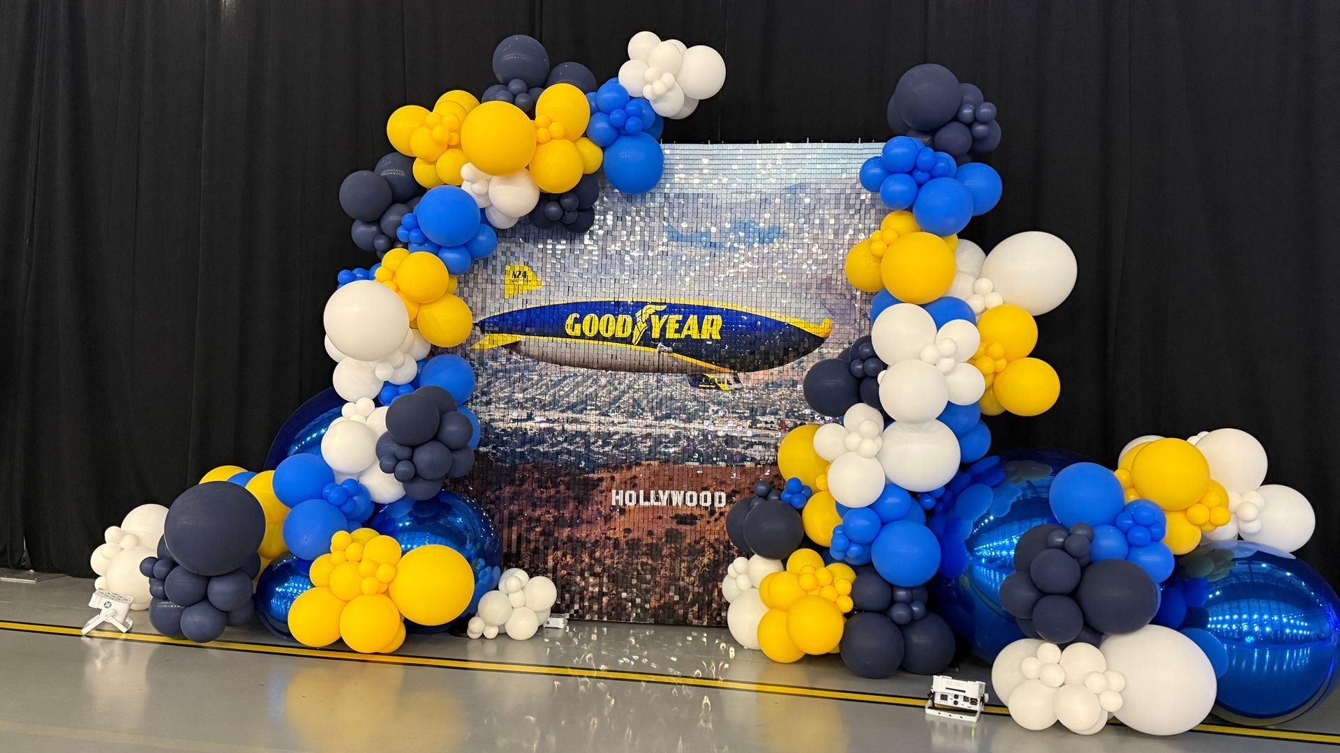 Goodyear Blimp photo backdrop with Shimmer wall framed by an organic balloon garland in blue, yellow, white, and navy with metallic blue accents.