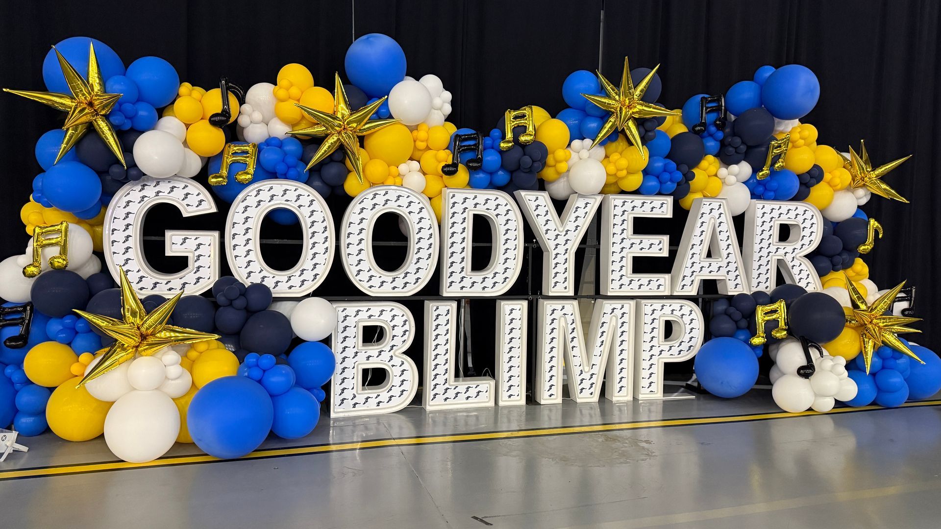 Double-stacked LED marquee letters spelling “GOODYEAR BLIMP,” surrounded by blue, yellow, white, and navy balloon décor with gold stars and musical notes.