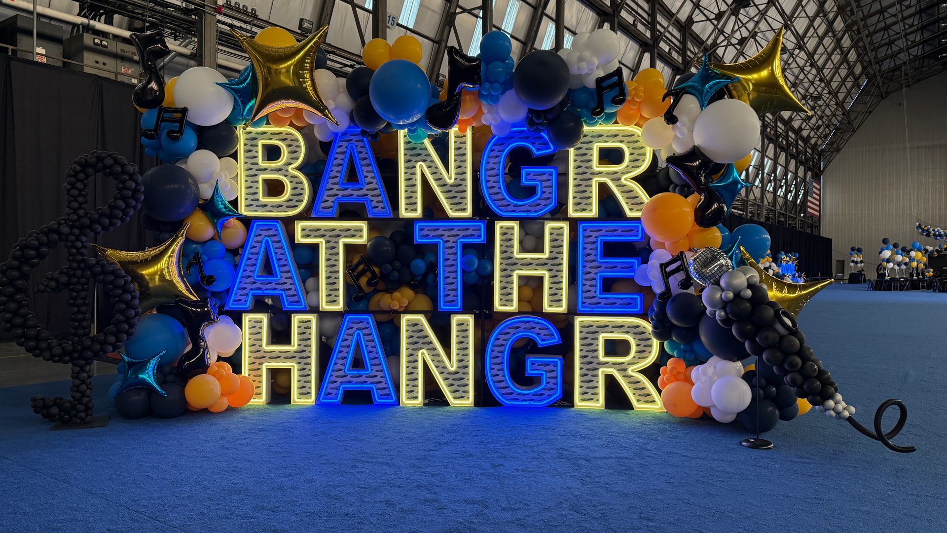 Triple-stacked blue and yellow LED marquee letters spelling “BANGR AT THE HANGR,” surrounded by balloon décor, metallic stars and musical notes inside an aircraft hangar.
