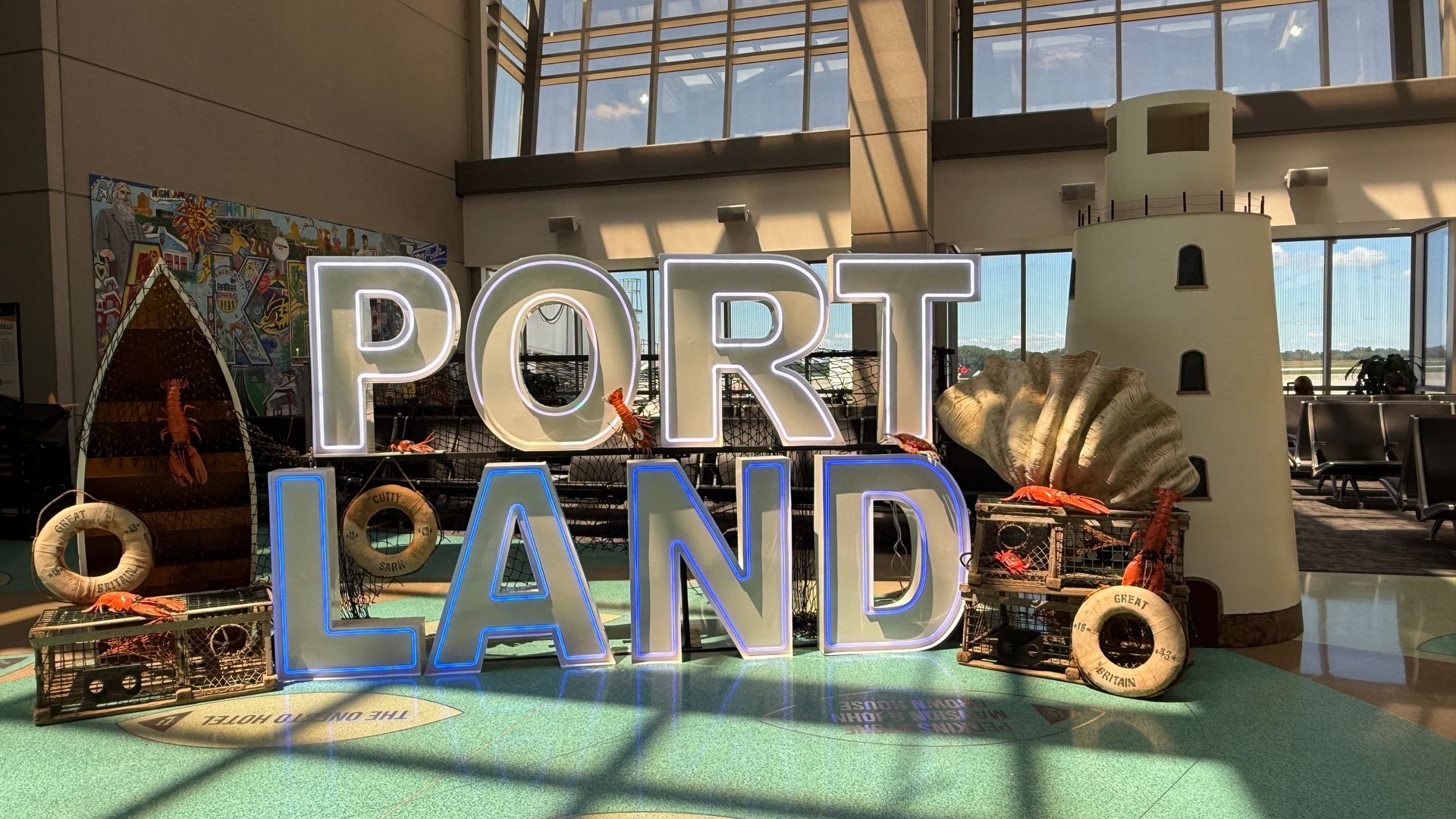 Illuminated “PORT LAND” LED marquee letters surrounded by lobster traps, buoys, seashells, and a lighthouse in a nautical-themed airport display.