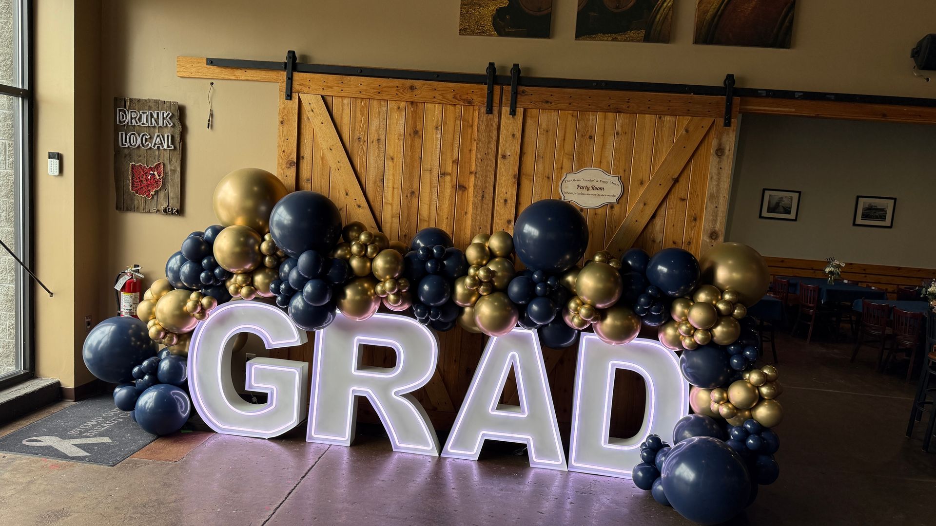 Illuminated GRAD marquee letters with a navy blue and metallic gold balloon garland at a graduation celebration.