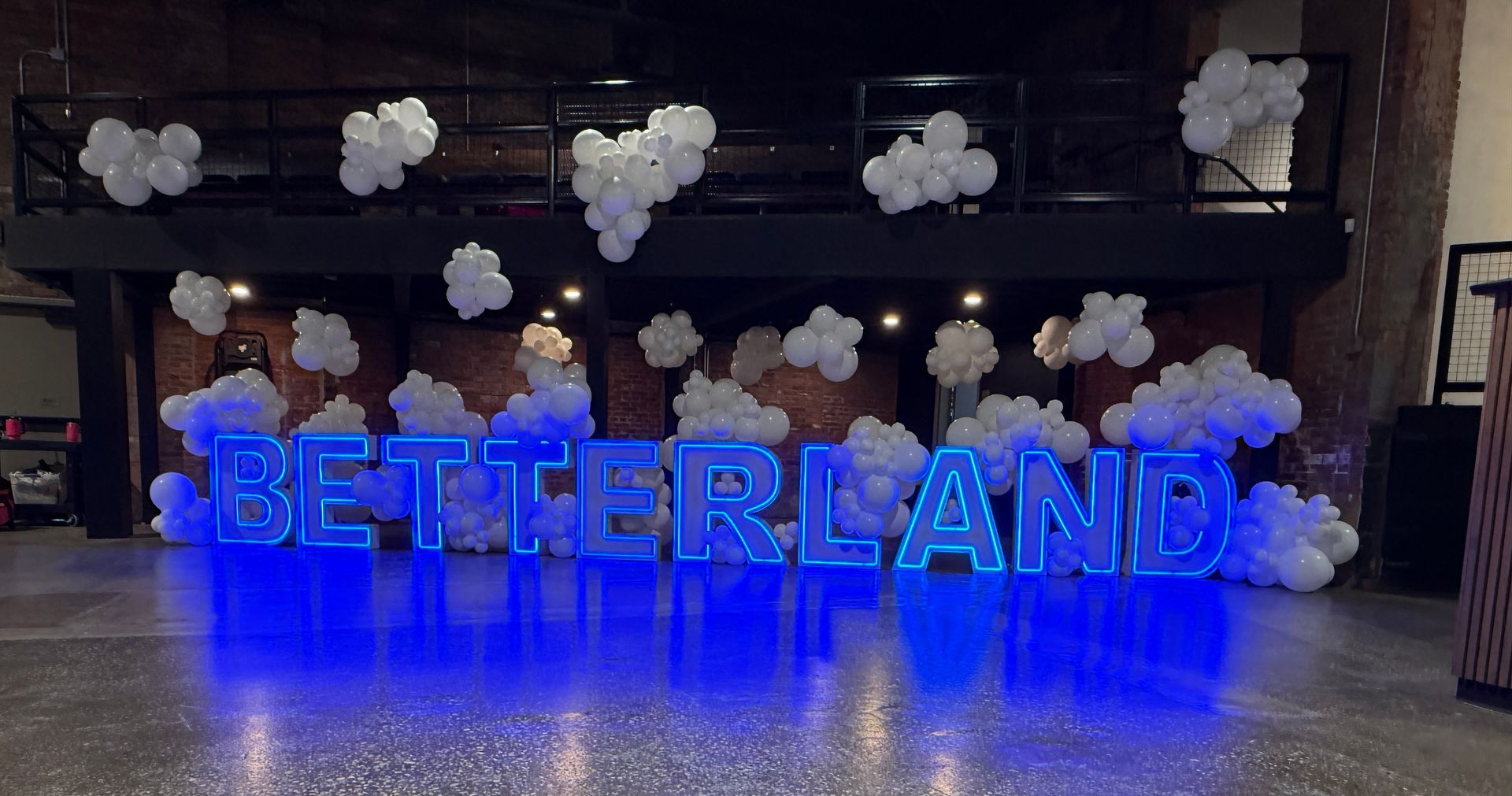 Blue LED marquee letters spelling “BETTERLAND,” surrounded by white balloon clouds inside an industrial-style event venue.