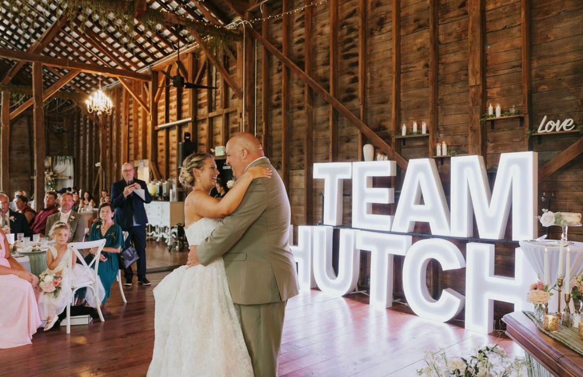 Bride and groom dance beside double-stacked, illuminated “TEAM HUTCH” marquee letters at their rustic barn wedding.