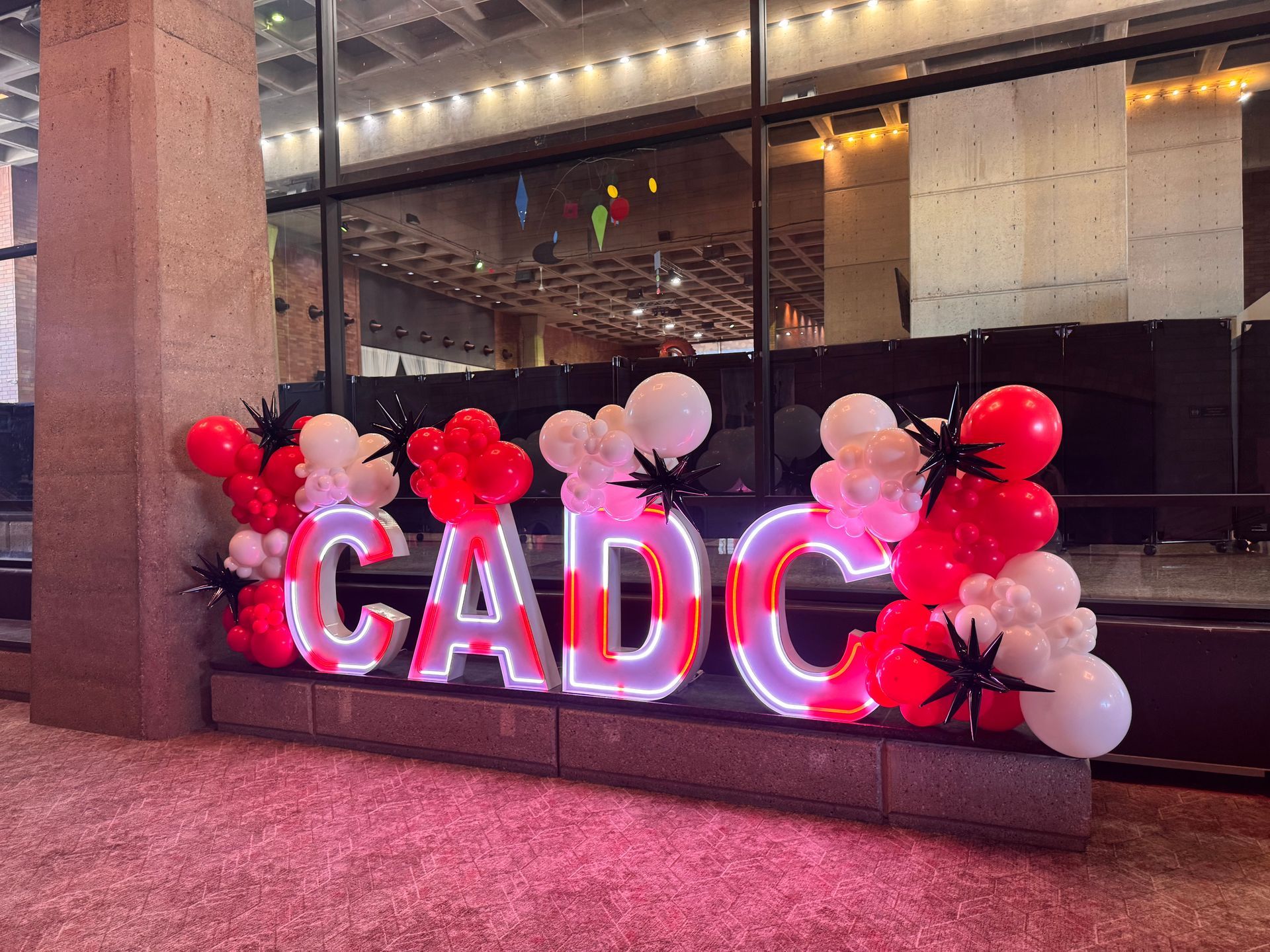 Illuminated “CADC” LED marquee letters with red and white organic balloon garlands and black starburst accents.