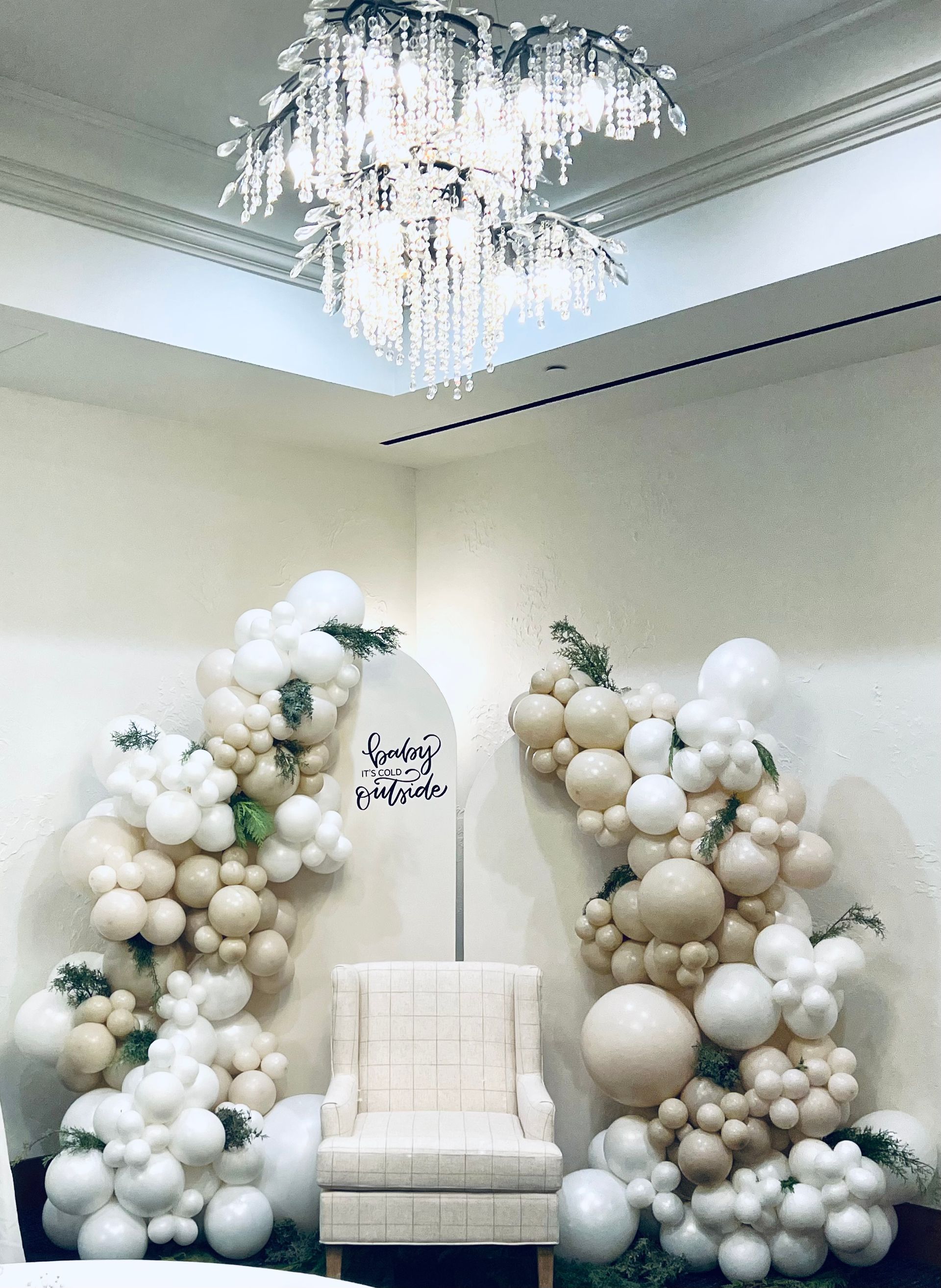 This elegant balloon backdrop features a stunning neutral-toned organic balloon arrangement in shades of white, ivory, and champagne, accented with greenery for a natural, winter-inspired touch.  The setup includes a softly curved arch sign with a phrase 