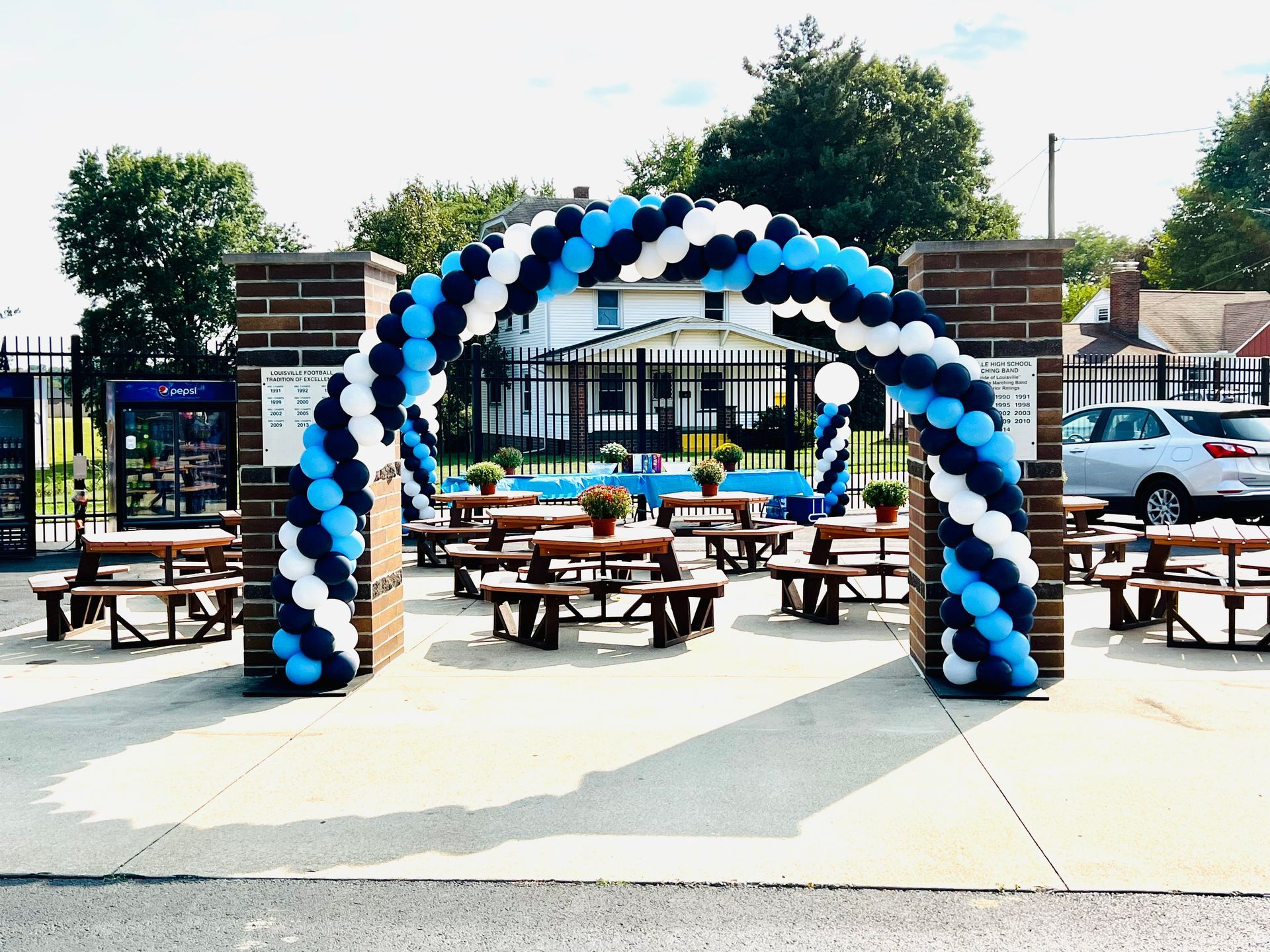 This classic spiral balloon arch features a bold mix of navy blue, pale blue, and white balloons, elegantly framing the brick entrance.  The arch enhances the outdoor setting, making it a perfect entrance statement for school events, sports games, grand openings, or community gatherings.  Its structured design adds a professional and festive touch, welcoming guests with style. 