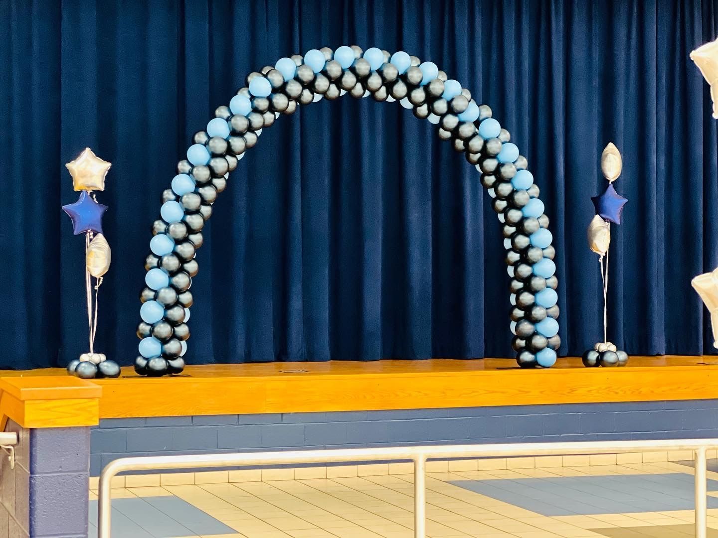 This is a classic spiral balloon arch featuring a combination of metallic silver, metallic navy blue, and pale blue.  The spiral pattern gives it a sleek, polished look, making it a perfect focal point for a stage setup.
Key Features:
Spiral Pattern: Alternating balloon colors create a structured and uniform designs.
Metallic Accents: The silver balloons add a touch of elegance and shimmer.
Stage Placement: The arch beautifully frames the stage, making it ideal for graduations, awards ceremonies, performances, or special presentations.
This setup is perfect for formal events such as school ceremonies, corporate galas, proms, or milestone celebrations, adding a sophisticated and eye-catching element to the stage. 