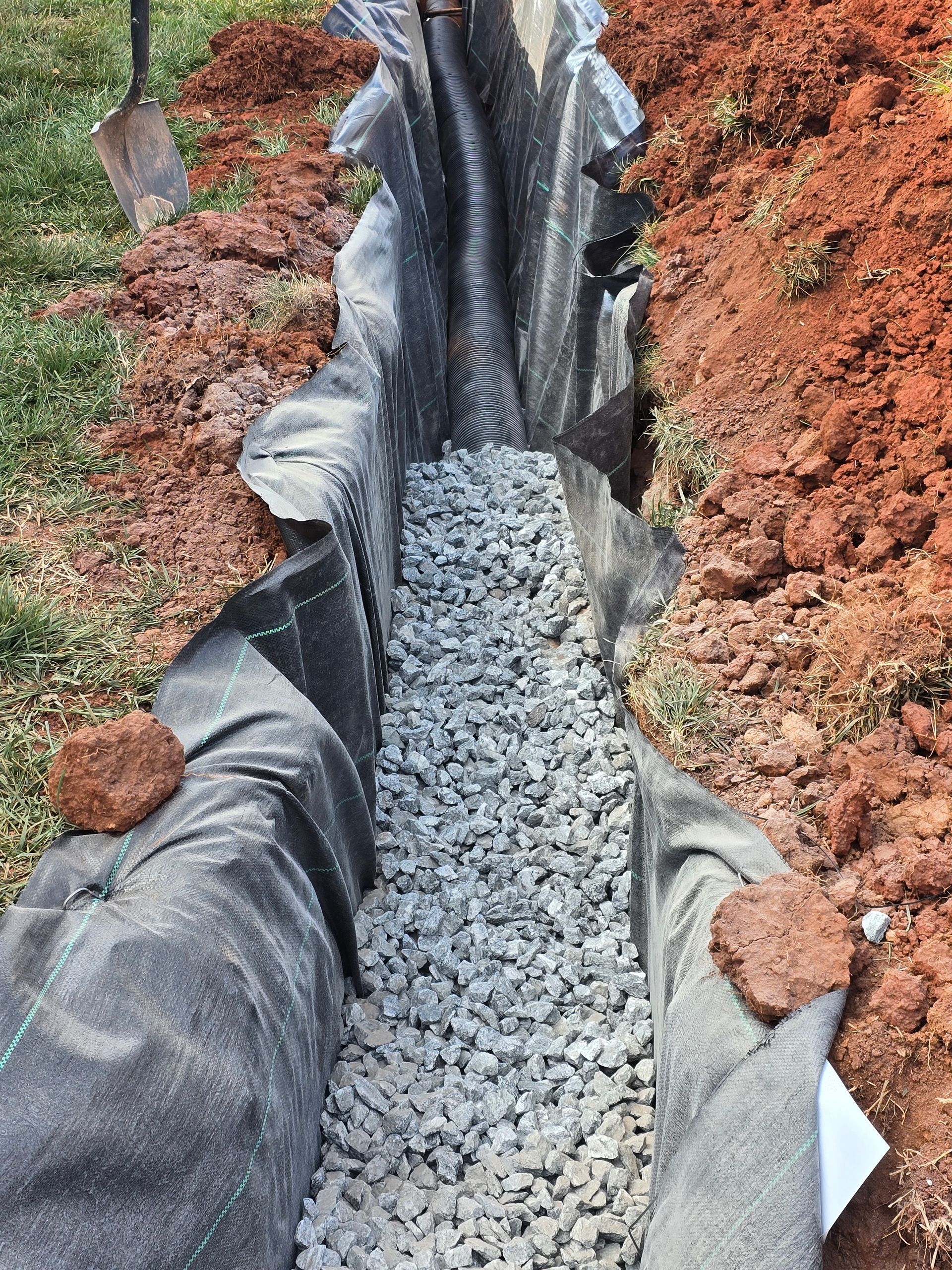 Gravel-lined drainage trench with black plastic edging through a dirt yard, holding gray stones.