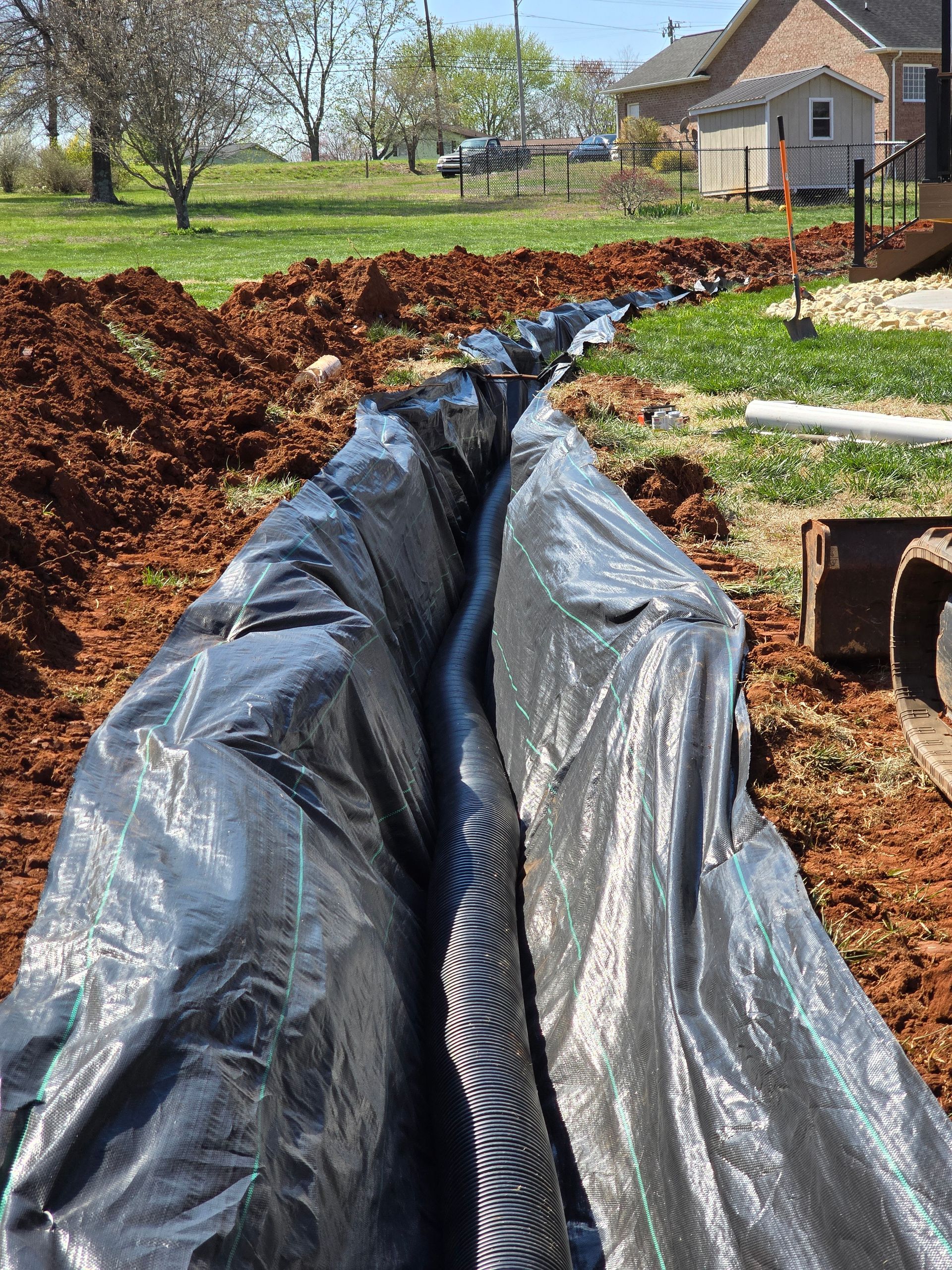 Black plastic-lined trench in a yard, with dirt piles, green grass, and a house in the background.