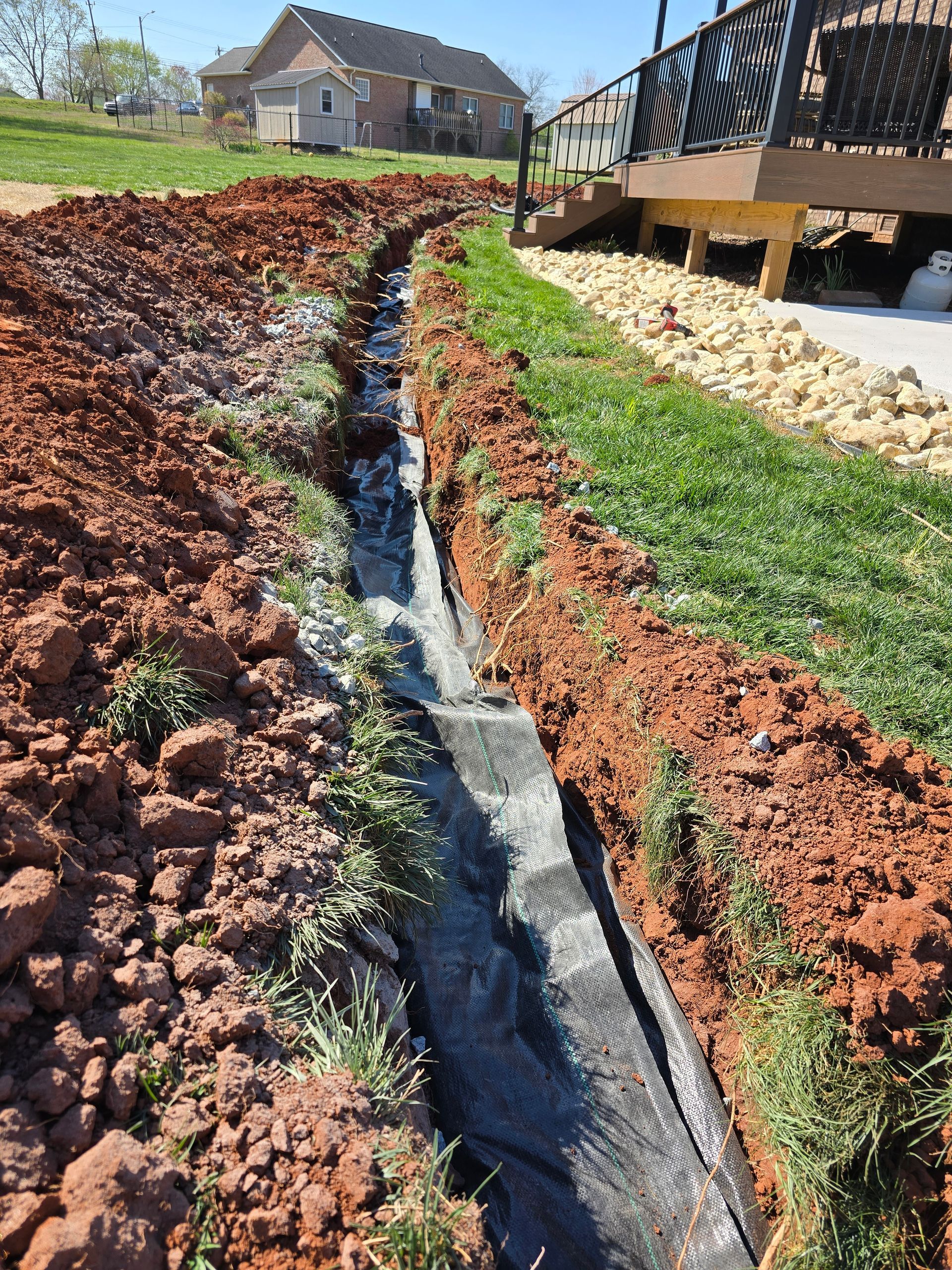 A narrow drainage trench lined with black fabric runs through fresh red dirt beside a raised deck and lawn.