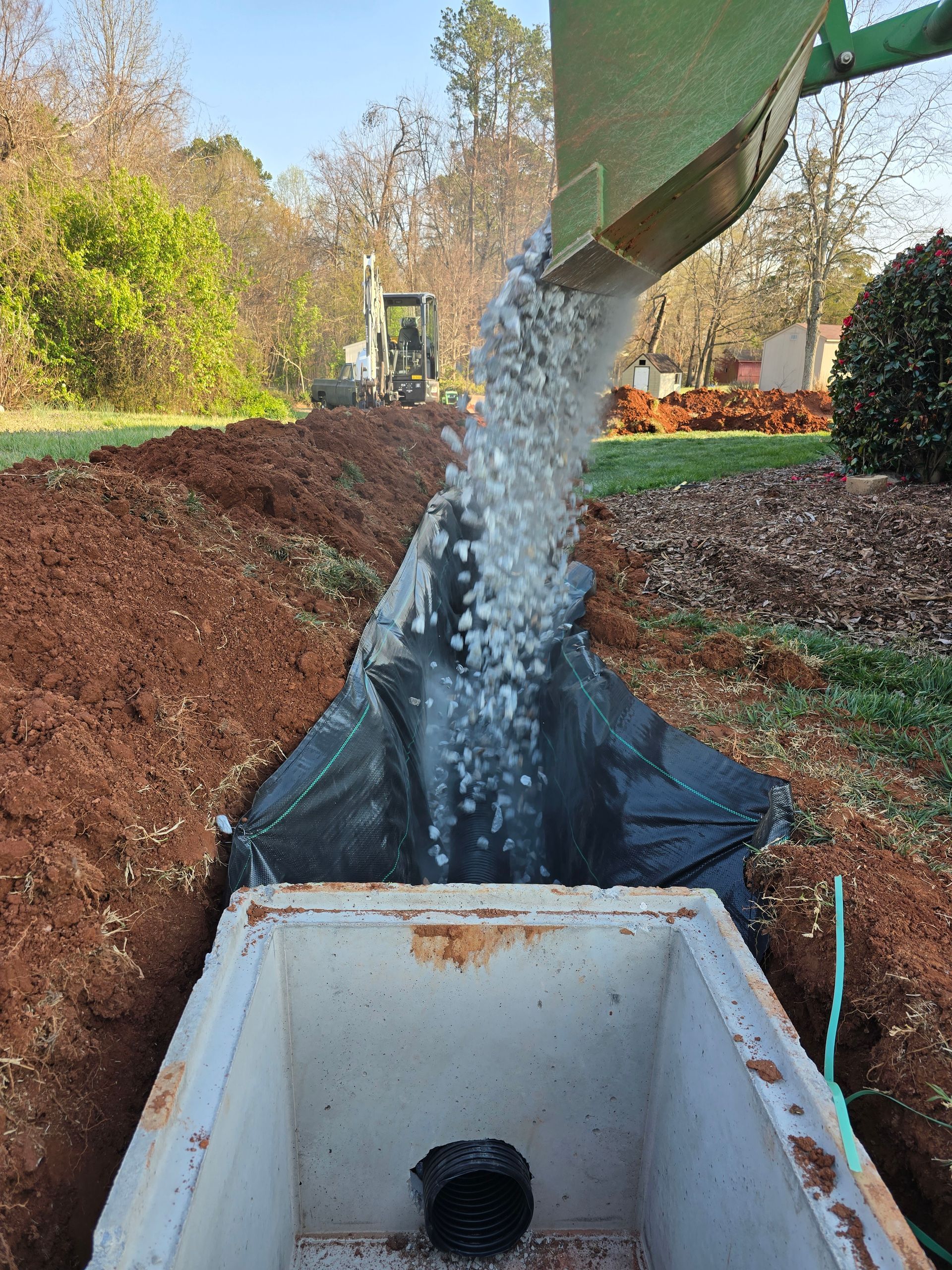Water pouring from a chute into a lined trench at a construction site with dirt piles and trees nearby