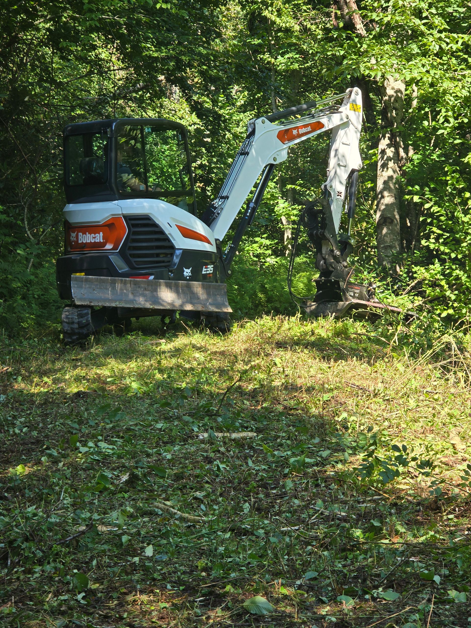 Small excavator clearing brush in a leafy forest clearing.