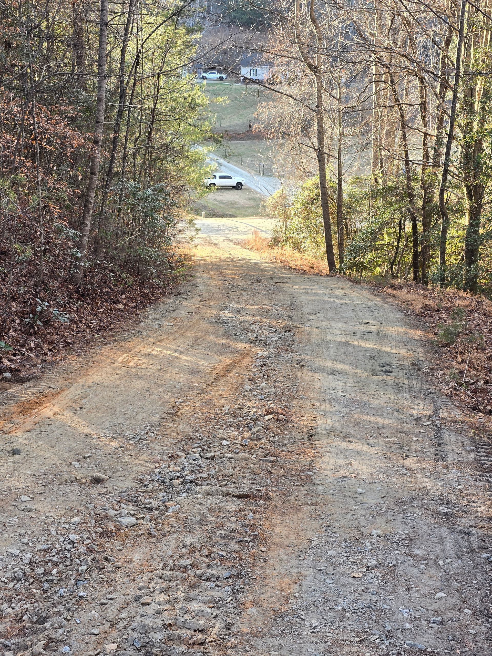 Gravel road leading downhill through trees toward a distant house in autumn.