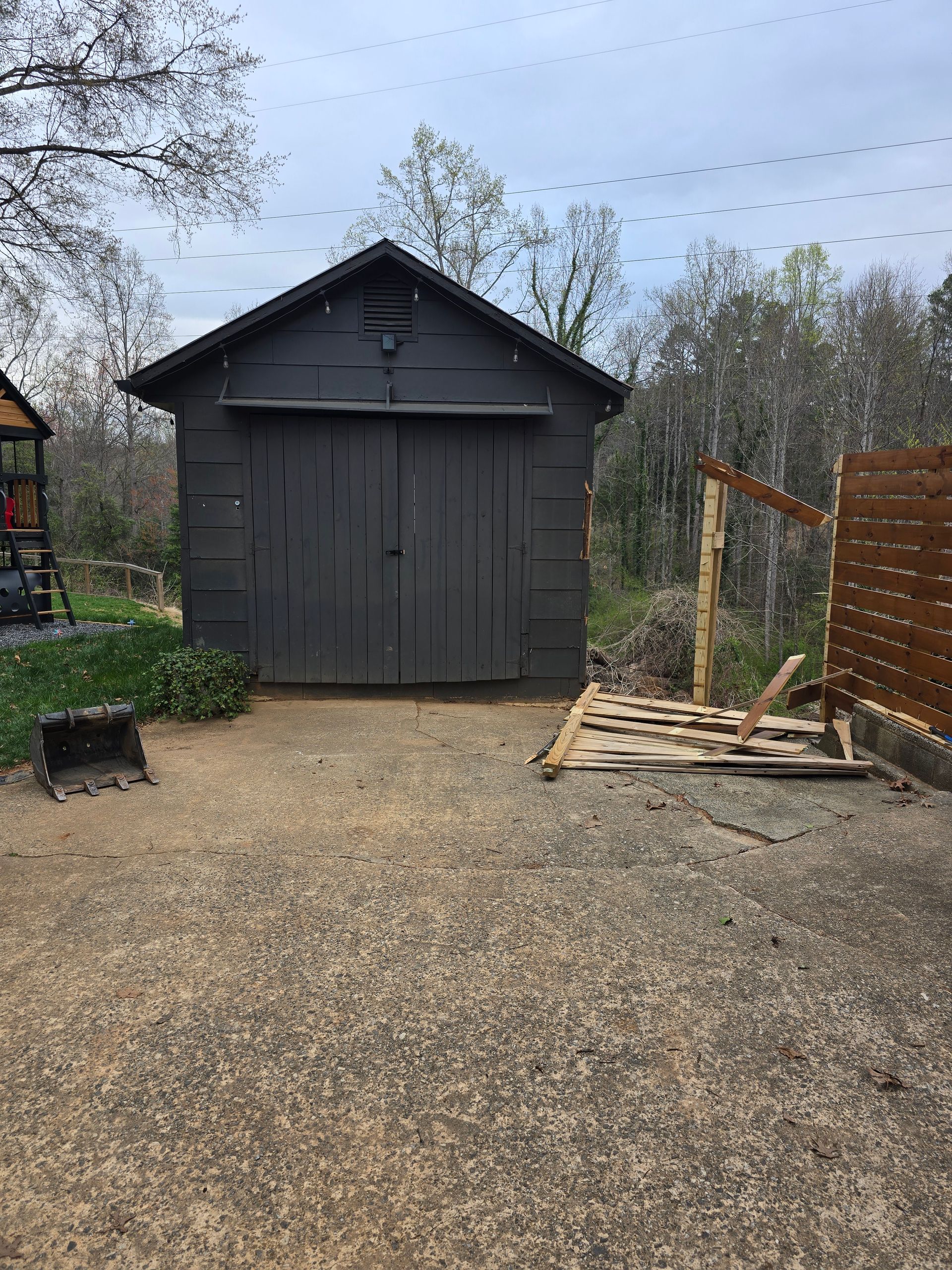 Dark gray shed in a gravel yard with a wooden pallet, fence, and trees in the background