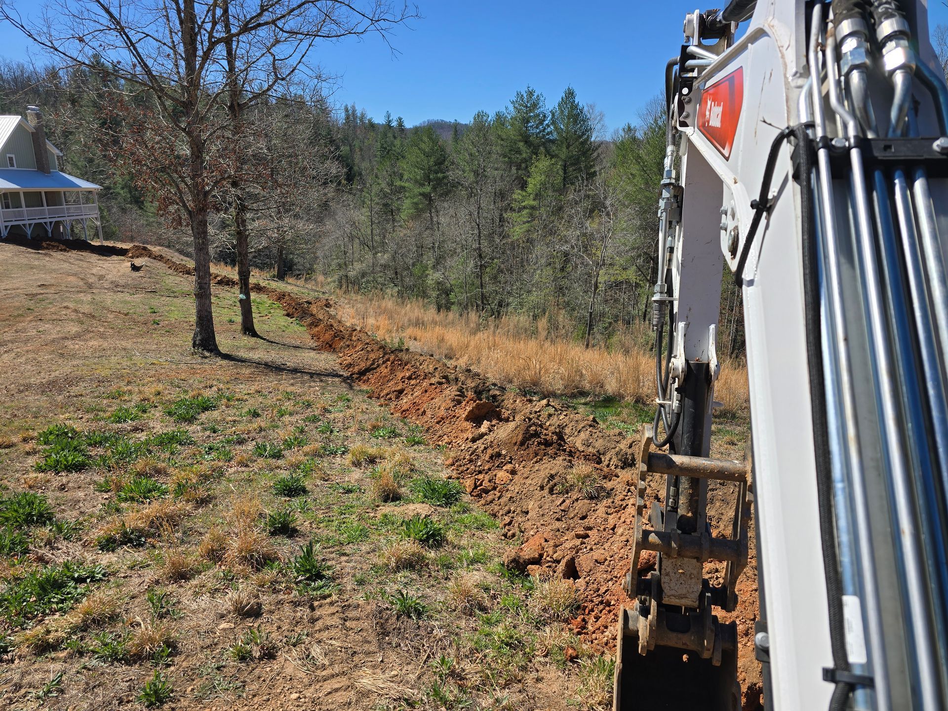 Excavator clearing a ditch beside a grassy field and trees under a blue sky