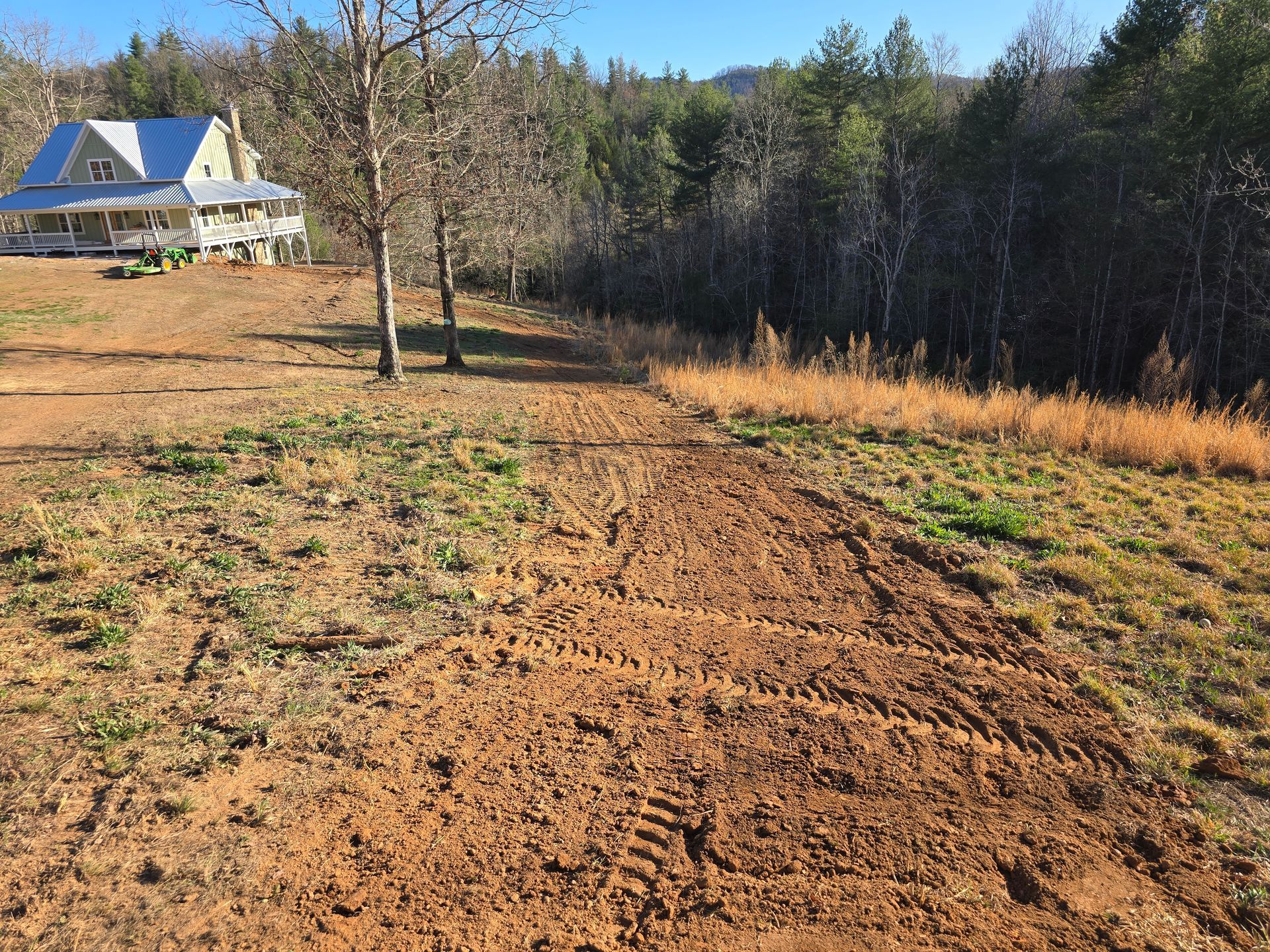 Rural yard with a dirt path, bare trees, grassy slopes, and a house with a blue roof in the background